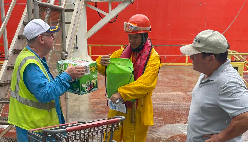 A volunteer handing a case of drinks and a welcome bag to a crew member on the dock